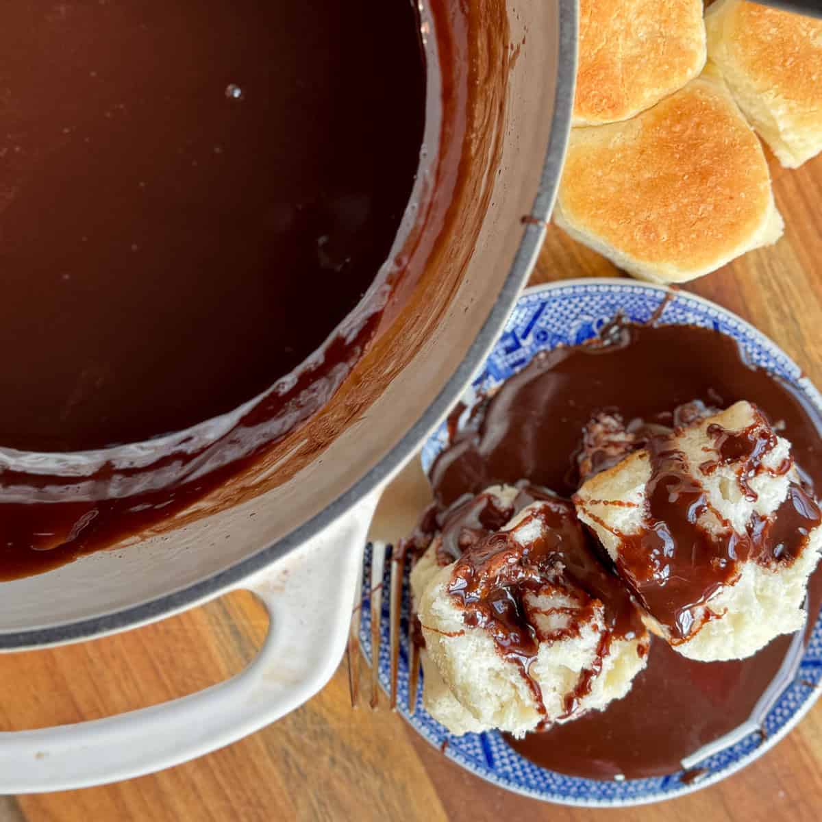 A pot of rich chocolate gravy sits on a wooden surface beside a blue-patterned plate with two biscuits covered in gravy. Several plain biscuits are arranged nearby. A fork rests near the plate.