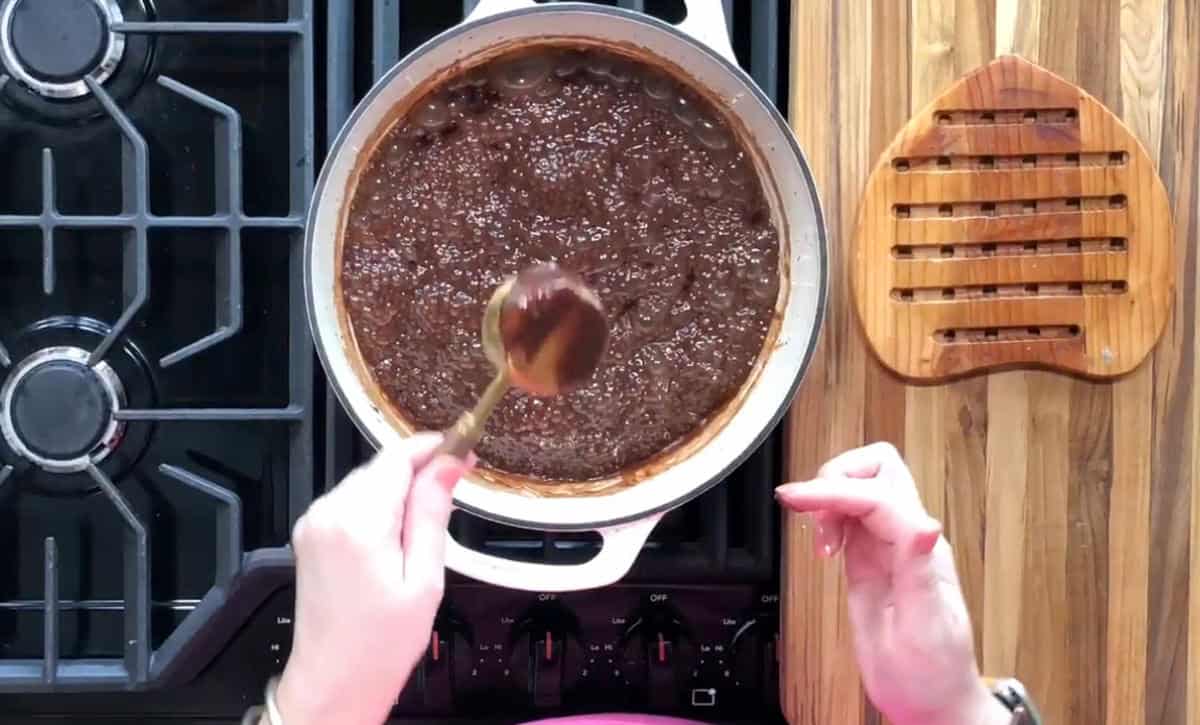 Top view of a person stirring a thick brown sauce in a pot on a stove. The hand holds a wooden spoon. A wooden trivet is placed on the countertop nearby. The stove's black surface has control knobs on the front.