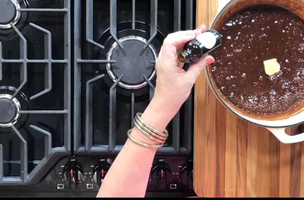 A person wearing bracelets holds a bottle over a pot filled with a dark liquid mixture and a pat of butter. The pot is on a wooden surface next to a gas stove with three visible burners and control knobs.