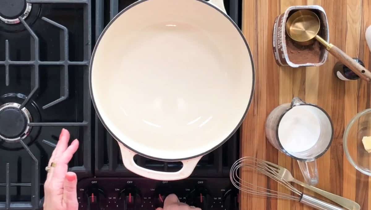 A person adjusts the heat of a stovetop burner beneath a white pot. Nearby, ingredients like milk, butter, a whisk, a container of cocoa powder with a wooden spoon and a measuring scoop, and a glass bowl are on a wooden countertop.
