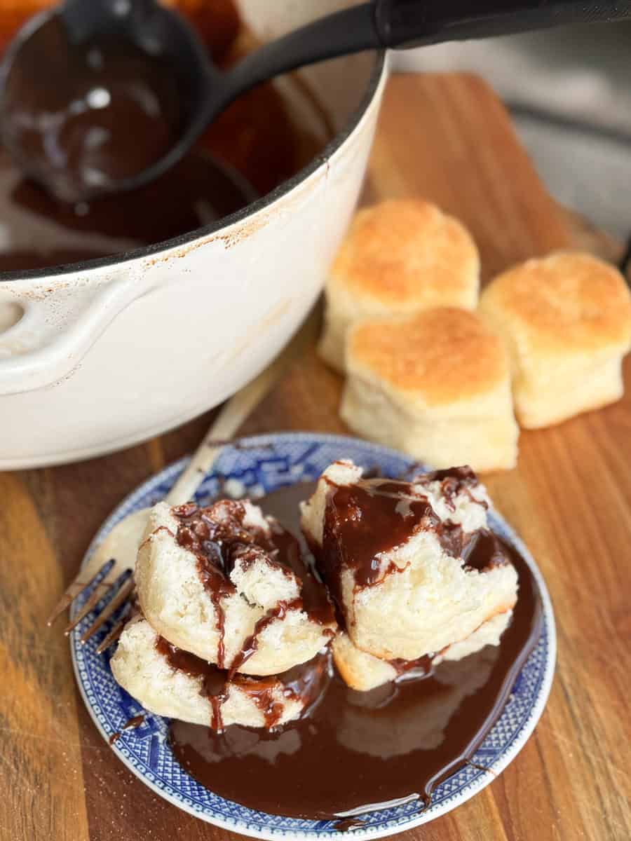 A plate with three biscuits topped with chocolate sauce sits on a wooden surface. A pot filled with chocolate sauce and a ladle is nearby. Three additional biscuits are in the background. A fork is placed beside the plate.