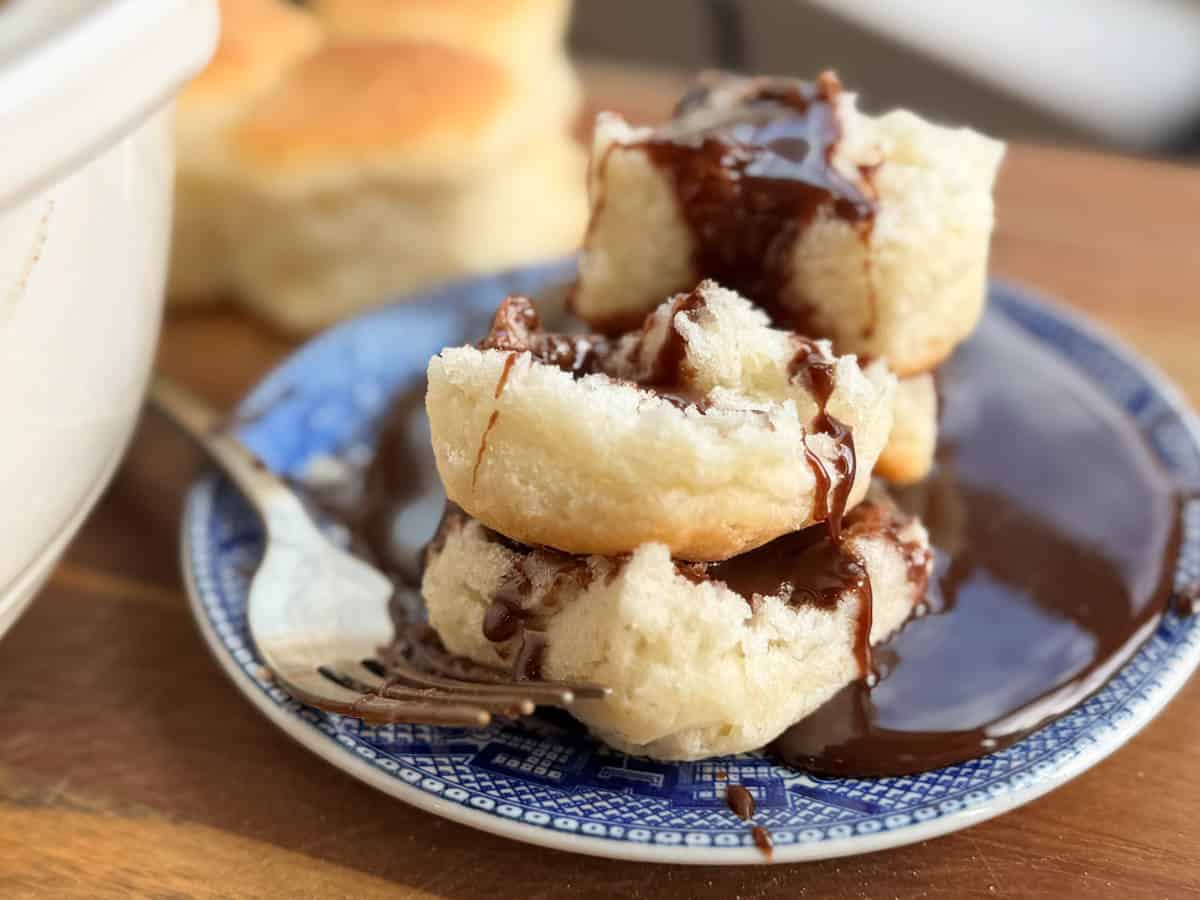 A stack of three biscuits is served on a blue and white plate, drizzled with a rich chocolate sauce. A fork rests on the plate beside the biscuits, and soft biscuits are visible in the background.
