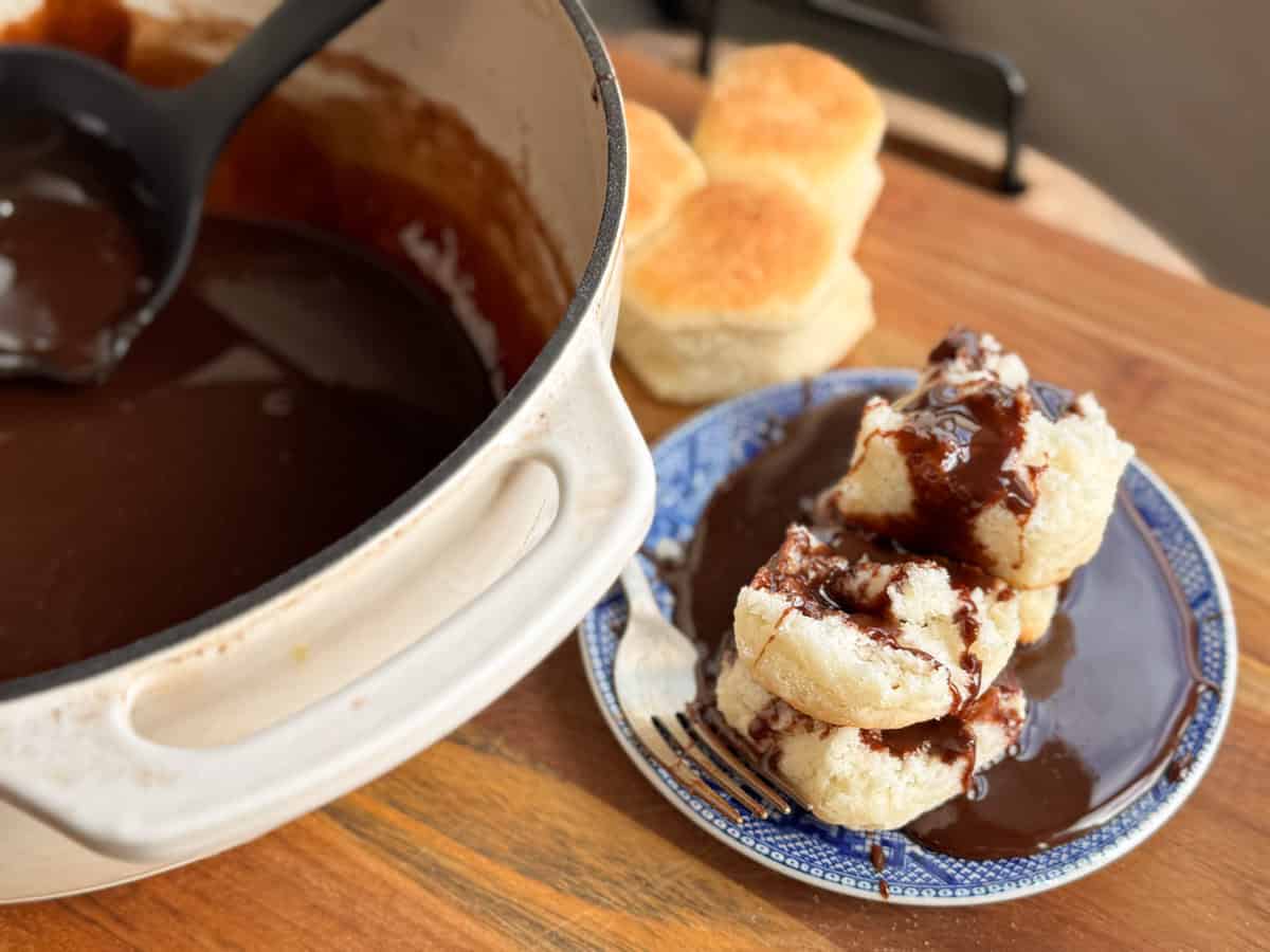 A white pot with chocolate gravy sits on a wooden surface alongside a plate with biscuits topped with the gravy. A ladle is resting on the pot's edge. Several plain biscuits are in the background.