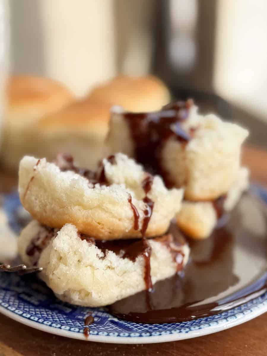 A stack of biscuits on a blue and white plate, topped with chocolate sauce. More biscuits are blurred in the background. The scene is brightly lit, highlighting the texture of the biscuits and the glossy drips of chocolate.