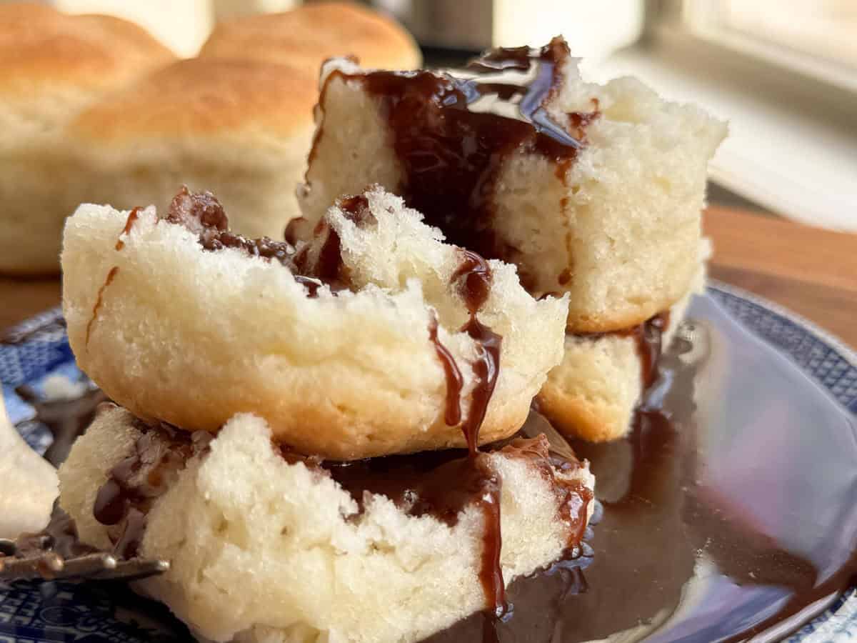 A close-up of a biscuit split in half, topped with melted chocolate sauce, placed on a blue-patterned plate. In the background, blurred biscuits are stacked on a wooden surface near a window.