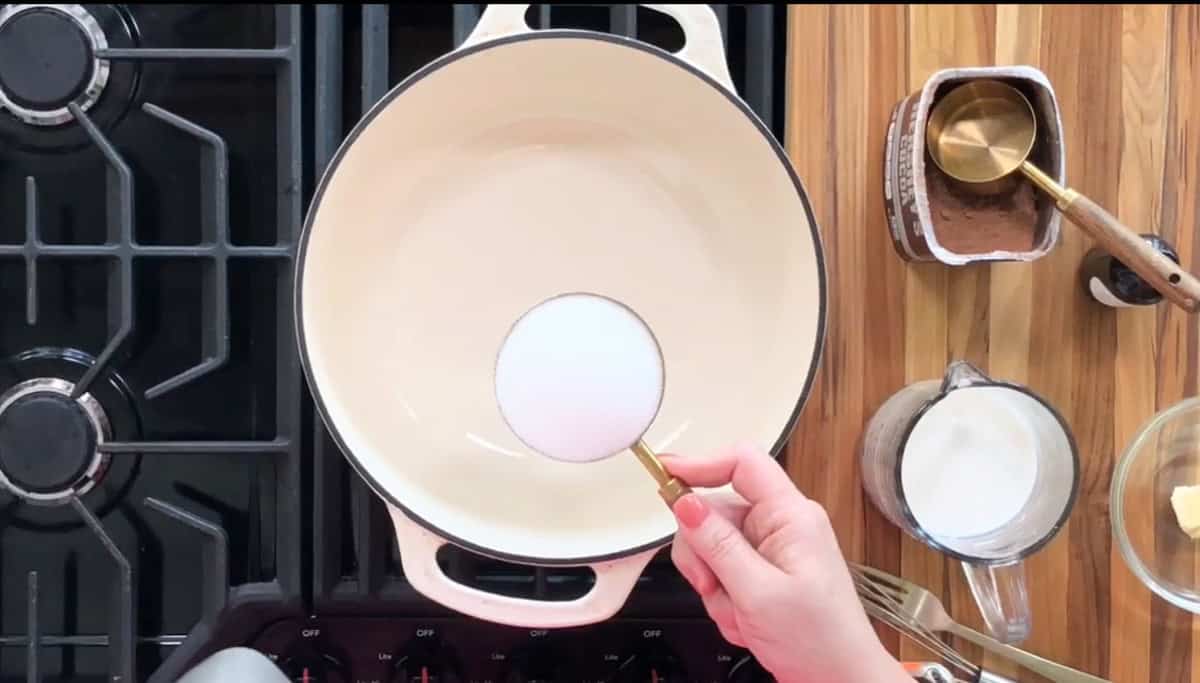 A hand holds a measuring cup of sugar over a cream-colored pot on a stovetop. To the right are containers with cocoa powder, milk, and butter on a wooden surface.