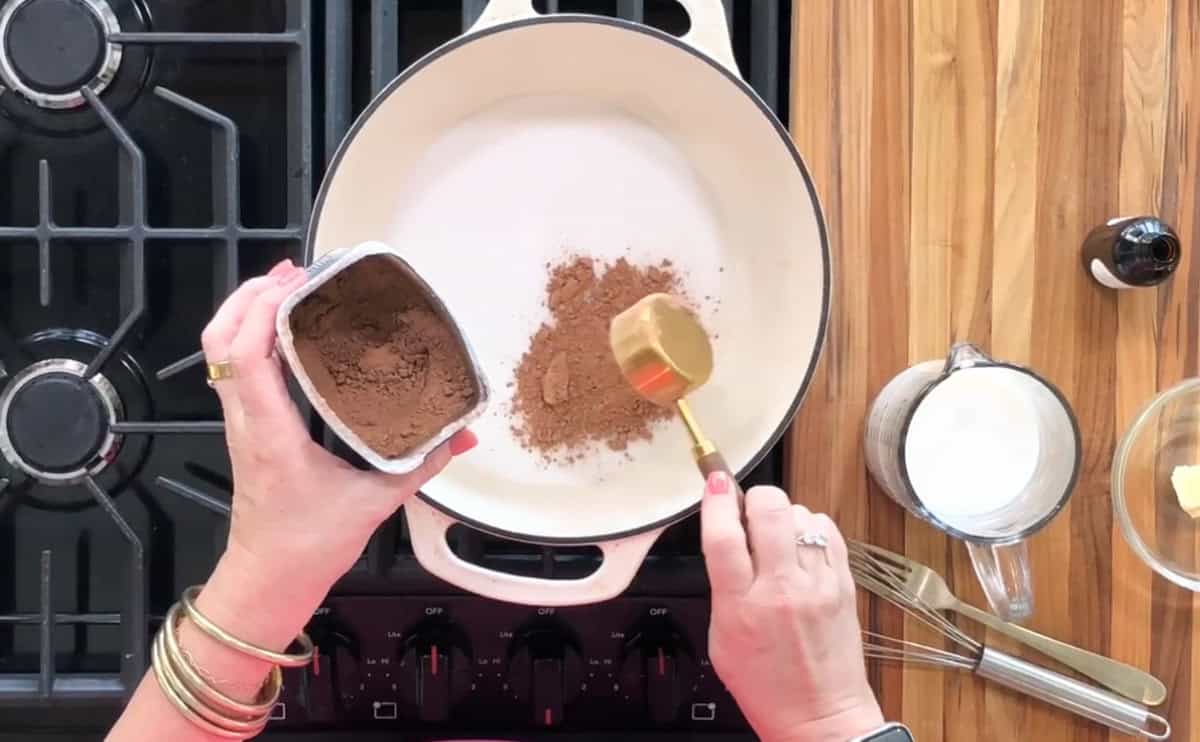 A person is adding a scoop of cocoa powder from a container into a white pot on a stovetop. To the right are a measuring cup with milk, a small bowl with butter, a whisk, and a bottle with dark liquid, all on a wooden counter.