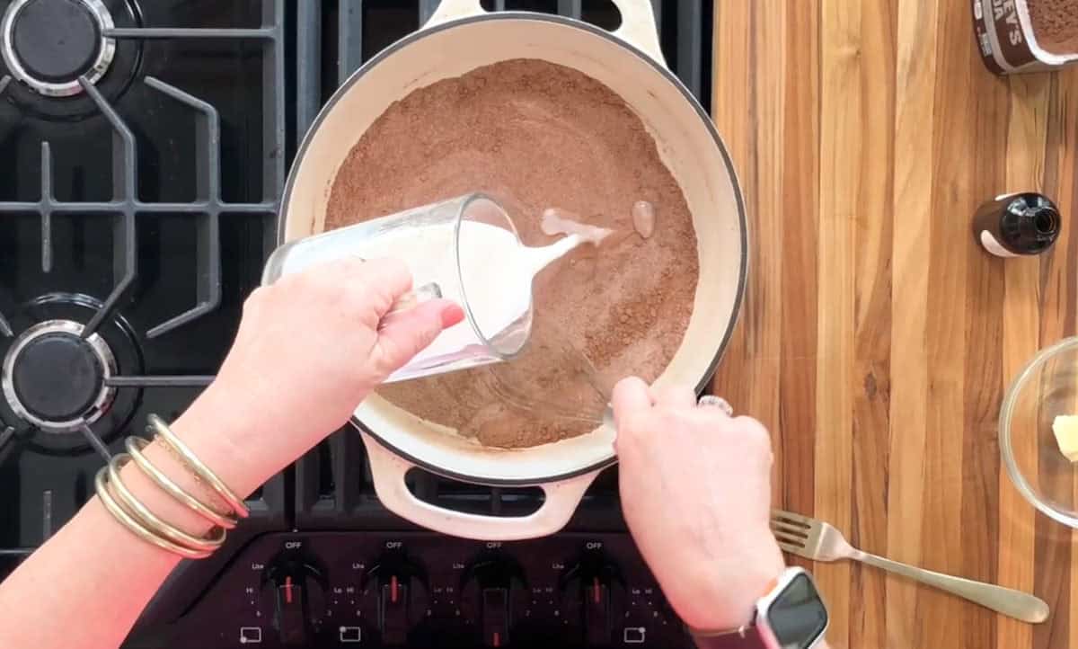 Person pouring milk from a glass into a pot containing brown powder on a stove. There is a wooden spoon in the pot, a dish with butter on the right, and a small bottle. The stove knobs and part of a wooden countertop are visible.