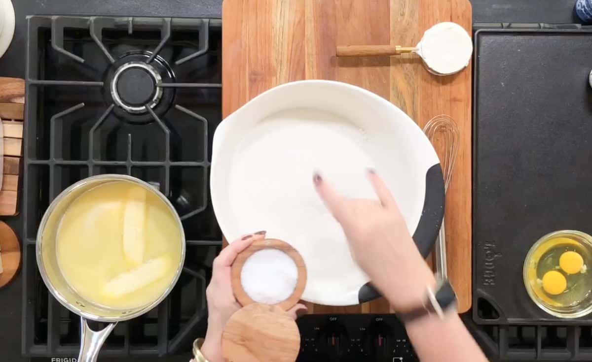 A person is preparing ingredients on a kitchen countertop. There is a stove with a saucepan containing melted butter, a bowl of sugar being measured, a small dish of salt, a whisk, a wooden spoon, a cutting board, and a bowl with two cracked eggs.