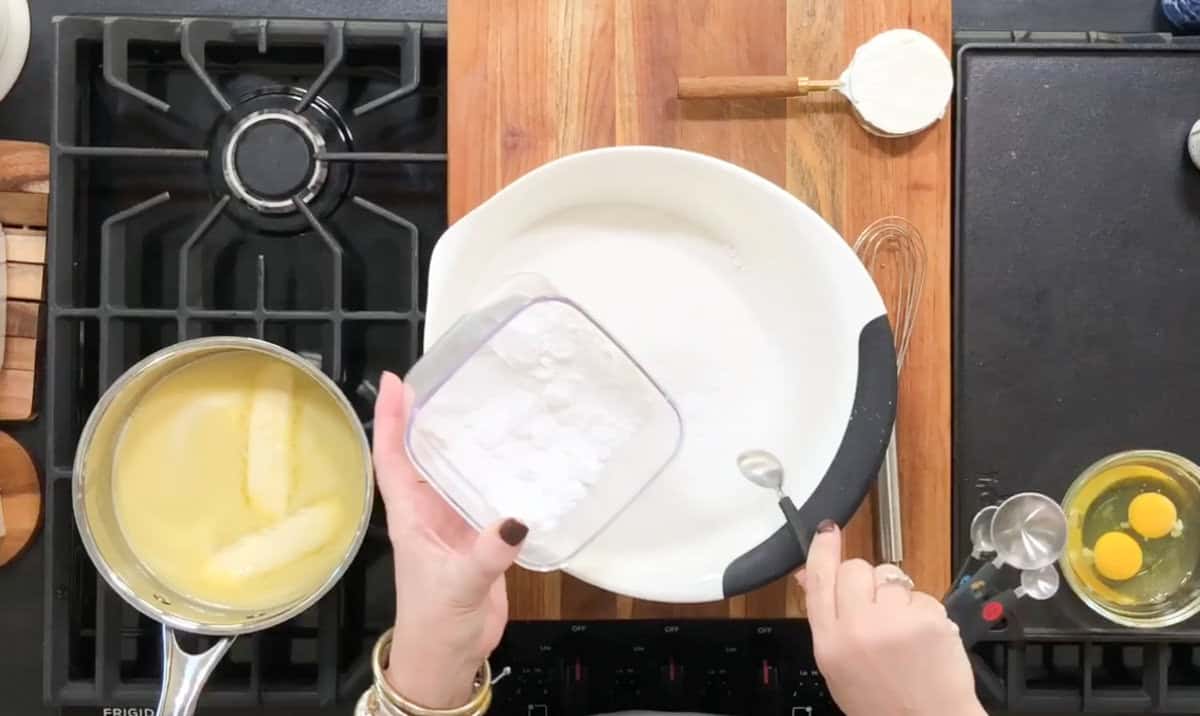 A person is preparing ingredients on a kitchen counter. A pot with melted butter is on the stove. Baking powder is being added to a white bowl. Two cracked eggs and a measuring cup with sour cream are nearby on the wooden board.