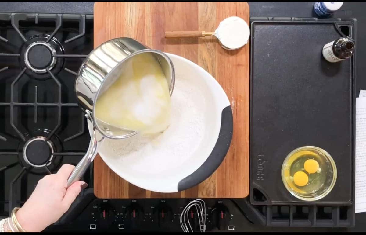 A person pours melted butter into a mixing bowl containing flour and sugar on a wooden cutting board. Nearby are a measuring cup with flour, a small glass bowl with two eggs, and a bottle of vanilla extract. The scene is set on a stovetop.