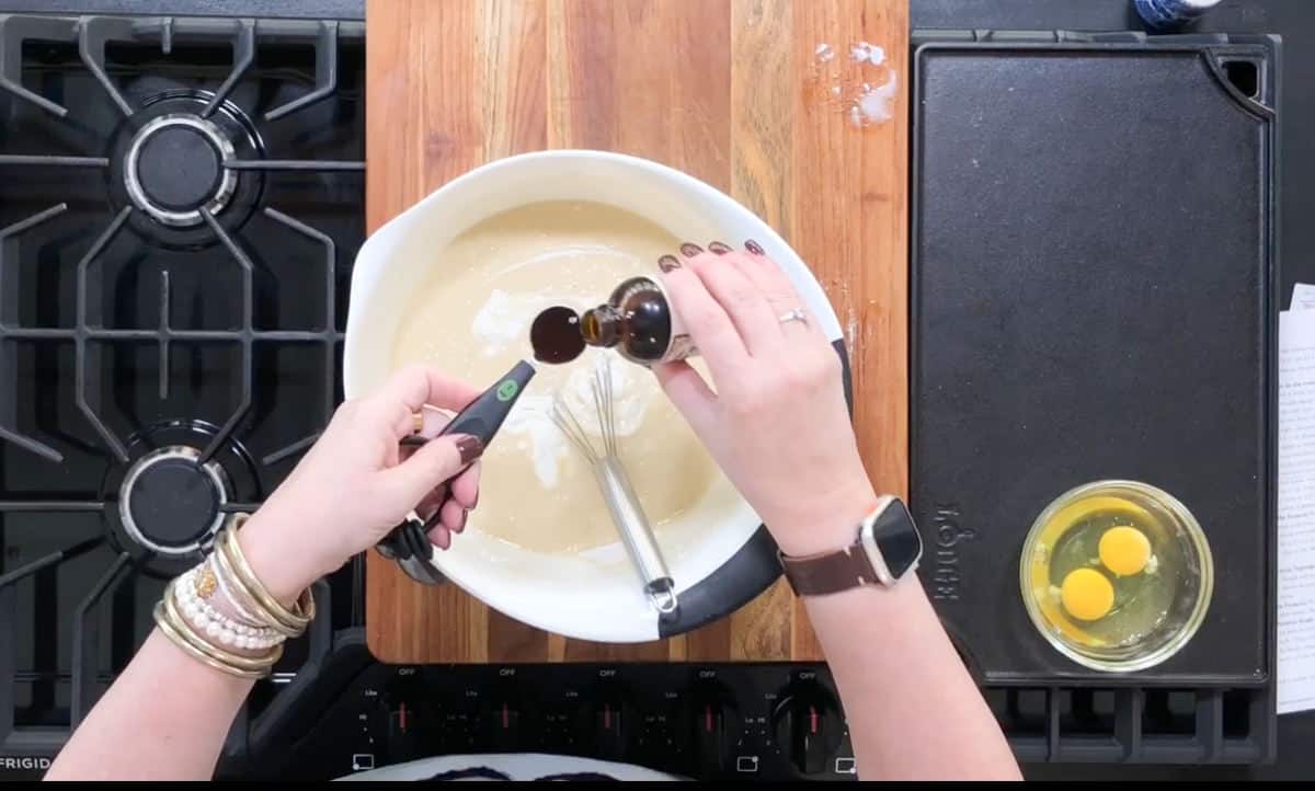 A person pouring vanilla extract into a mixing bowl filled with batter. The bowl is on a wooden cutting board between two stovetop burners. To the right, there is a bowl containing two eggs. A whisk and a spatula are in the mixing bowl.