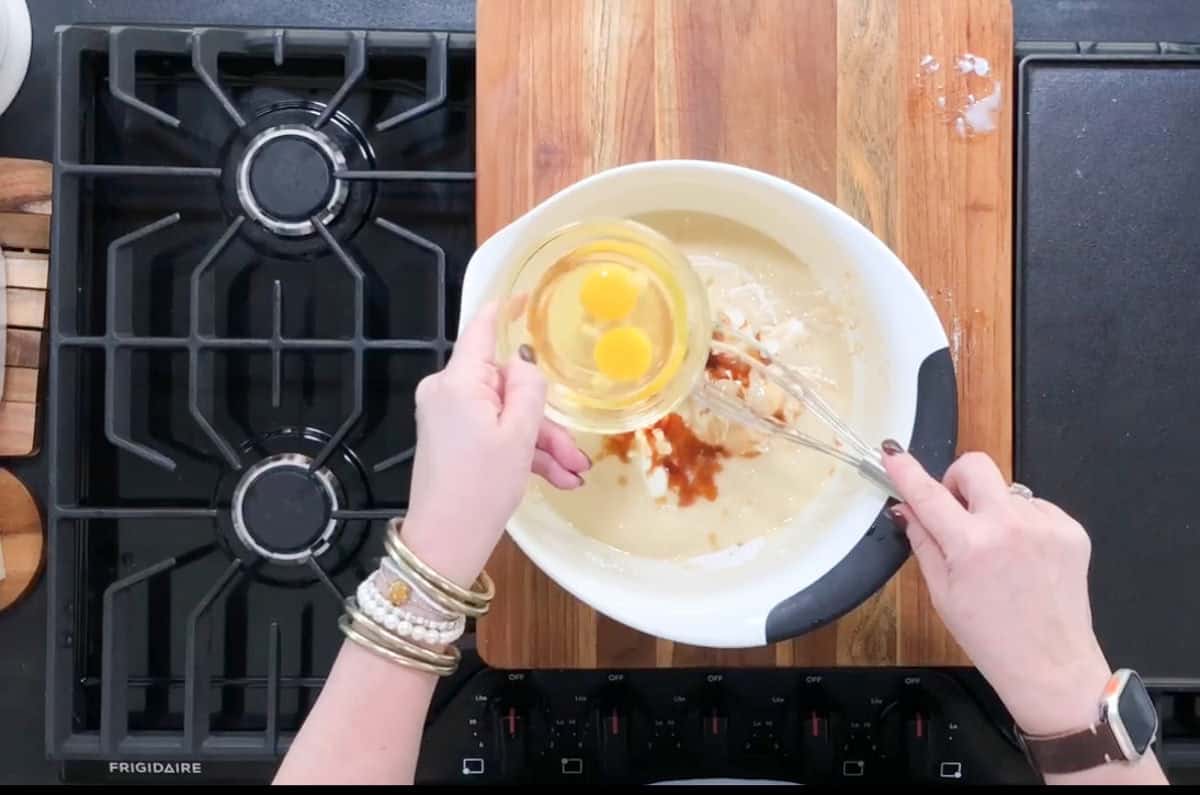 Hands are seen holding a bowl with two cracked eggs above a mixing bowl containing batter. A whisk is in the batter. The setup is on a wooden board beside a black stove with a Frigidaire logo. An Apple Watch is visible on the left wrist.