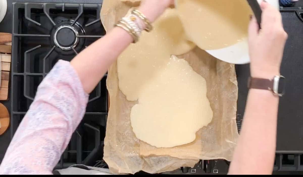 A person is pouring a creamy batter from a bowl into a parchment-lined baking tray. The tray is on a black stovetop, and the person is wearing a pinkish long-sleeve shirt and a watch. Several bangles are on their wrist.