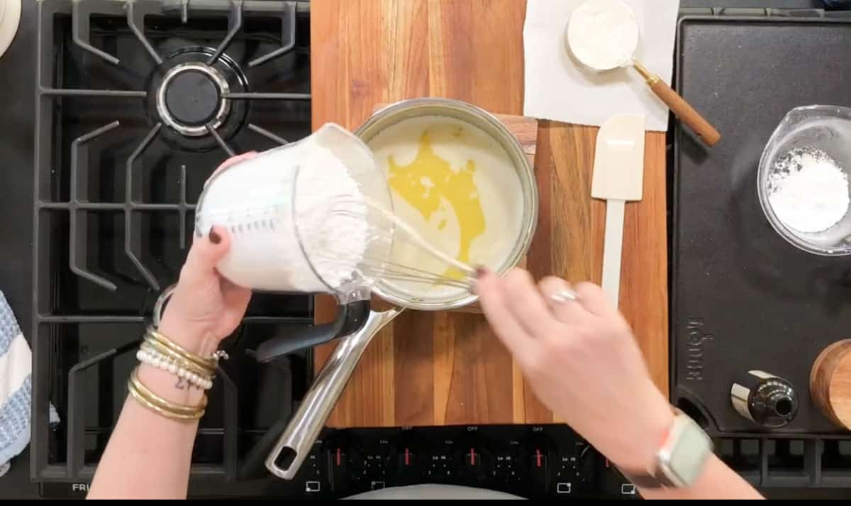 A person is stirring a saucepan containing a yellow liquid on a stove. They are holding a measuring cup filled with white powder, pouring it into the pan. The kitchen counter has a spatula, a bowl, and a towel nearby.