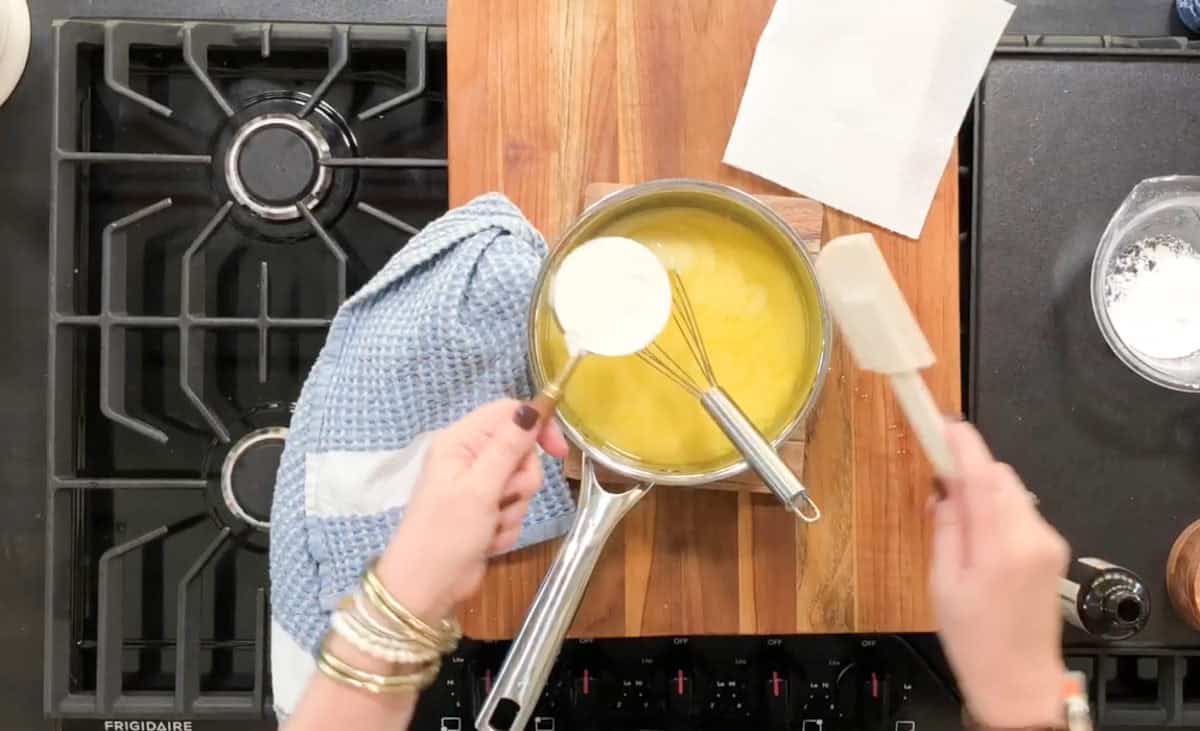 Overhead view of a person cooking on a stove. They are holding a metal whisk and adding a cup of flour to a saucepan with a yellow mixture. A blue towel is on the left, and a paper towel on the right.