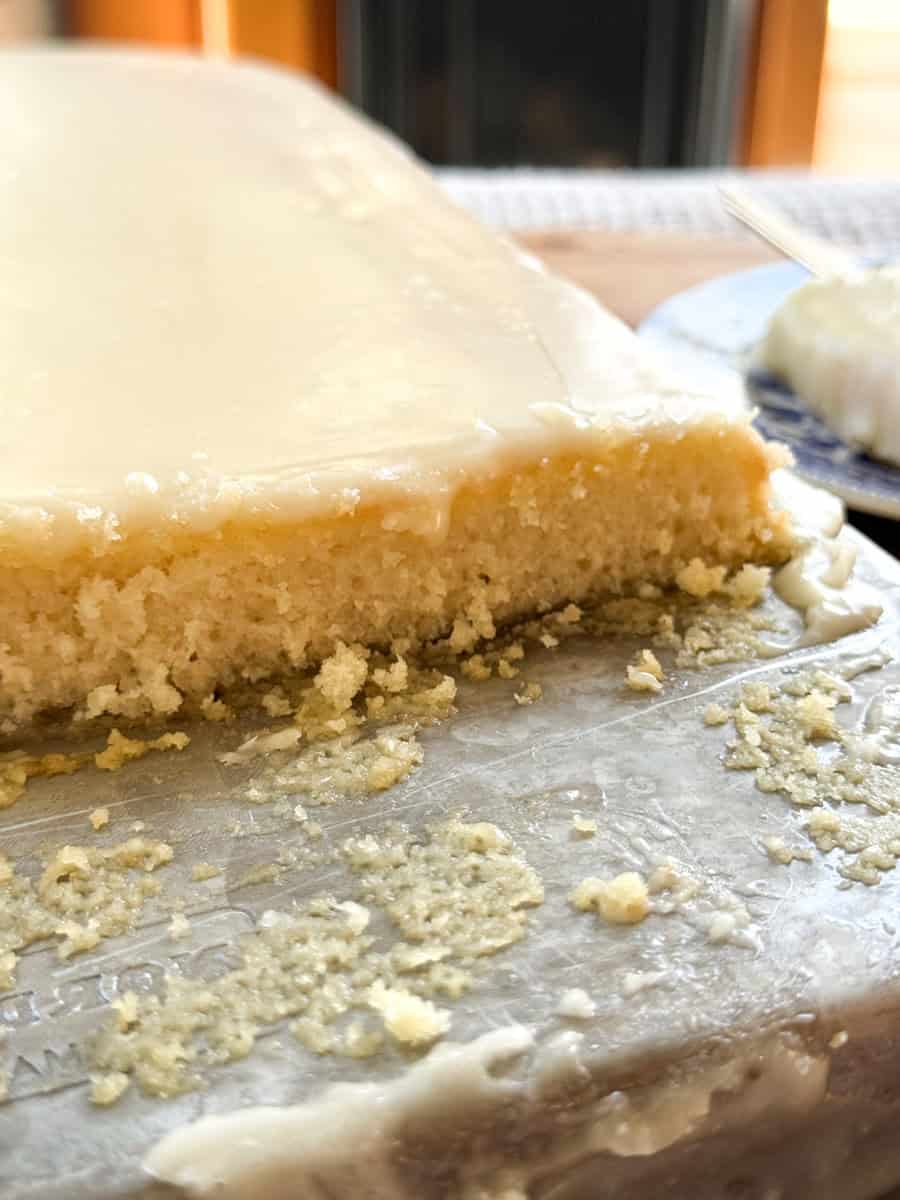A close-up of a vanilla cake topped with a layer of cream frosting. A piece has been cut from the corner, revealing the moist texture of the cake. Crumbs are scattered on the surface around the cake. A partially visible plate is in the background.