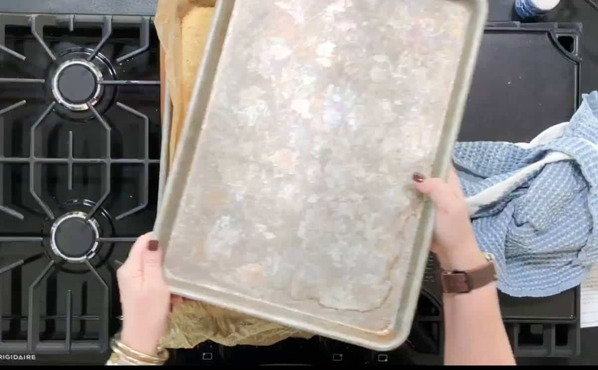 A person with painted nails holds a used baking sheet above a stovetop. A blue kitchen towel is visible on the right side. The oven's brand name, Frigidaire, is partially visible in the bottom left corner.
