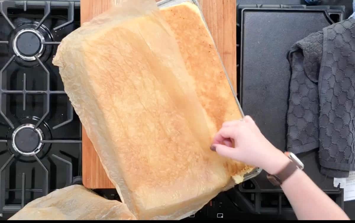 A person is peeling parchment paper off a large rectangular sponge cake on a wooden board. The board is on a stove, with two burners and a black cloth nearby. The person's hand is visible, wearing a watch.