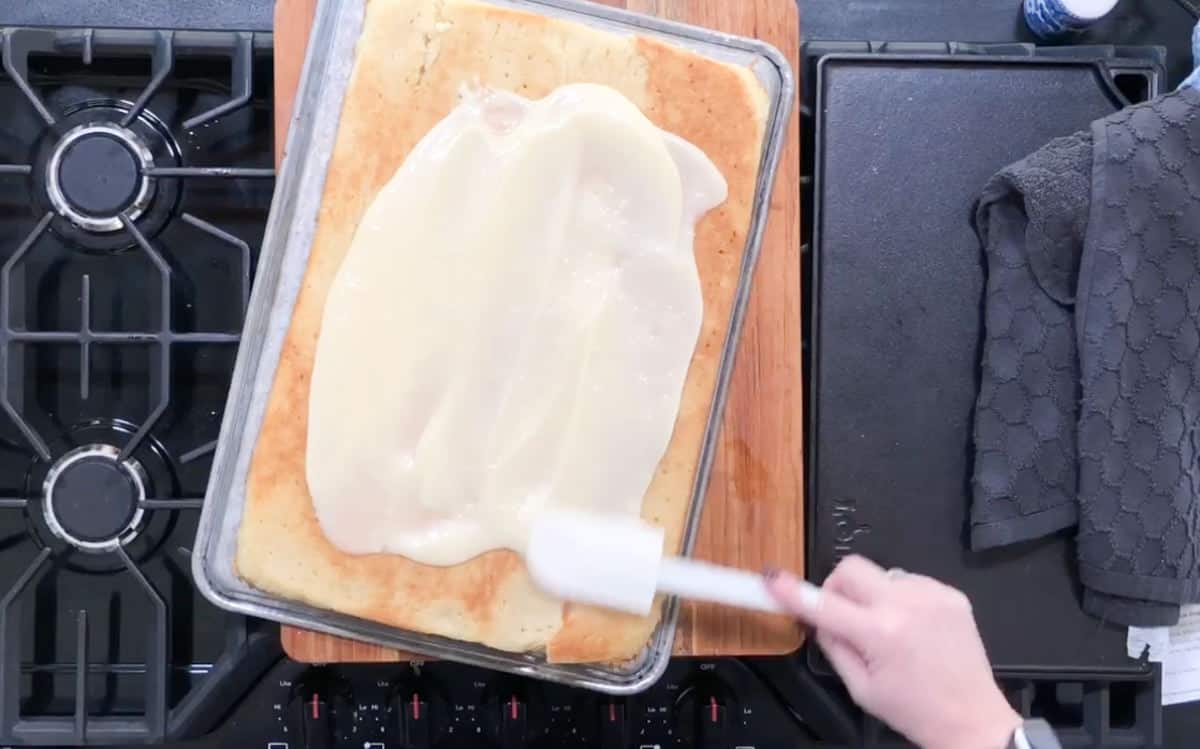 A person spreads a thick layer of white frosting over a large rectangular cake on a baking tray. The cake is positioned on a stovetop, with a black dish towel nearby. The person's hand holds a spatula to evenly distribute the frosting.