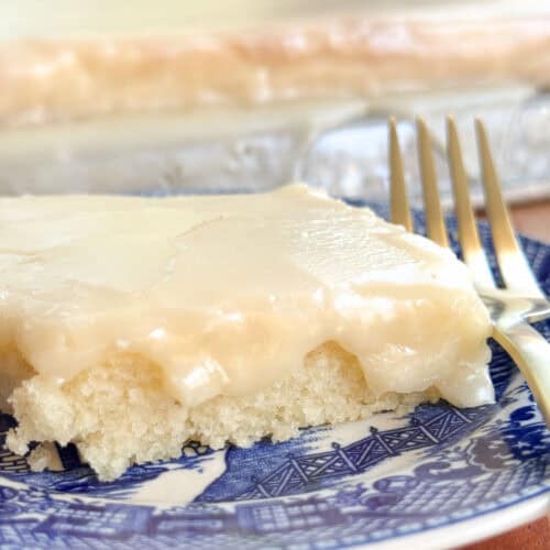 A piece of frosted white cake sits on a blue patterned plate with a metal fork beside it. The cake has a smooth, creamy icing and appears to be part of a larger batch in the background.