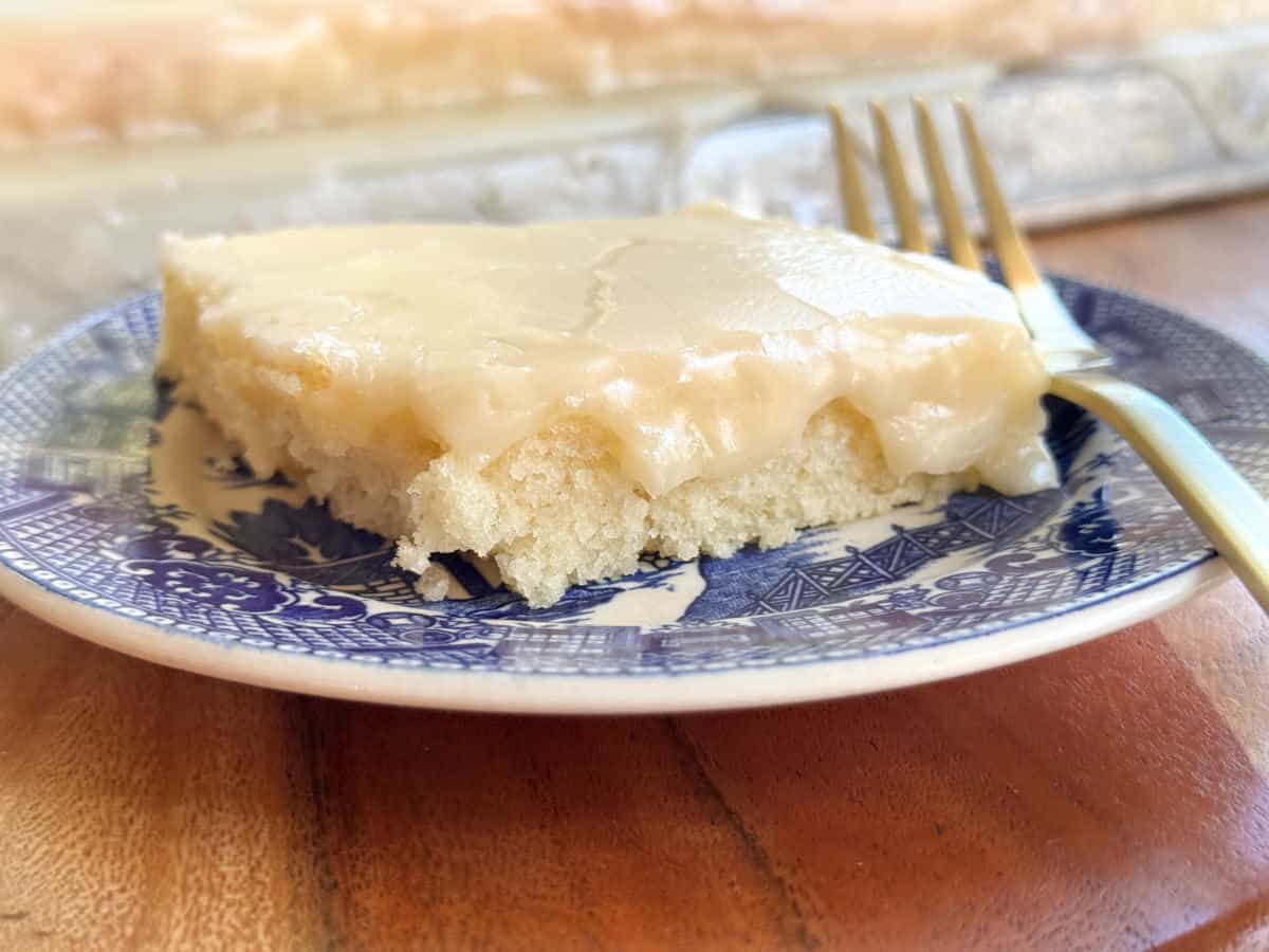 A slice of white cake with glossy icing on a blue and white plate. A fork rests beside the cake. The cake is on a wooden table with more cake visible in the background.