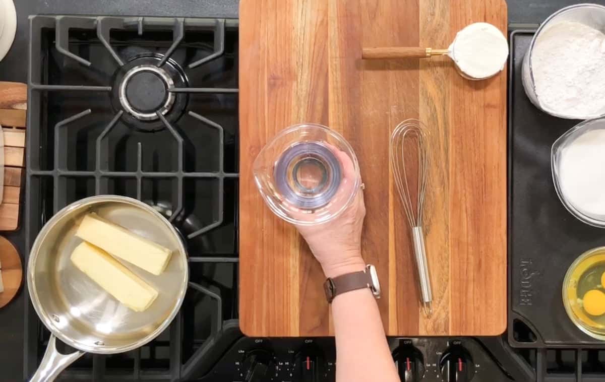 A person's hand holding a clear mixing bowl over a wooden cutting board. Surrounding items include a whisk, flour, sugar, eggs, and a saucepan with butter on an active stovetop.