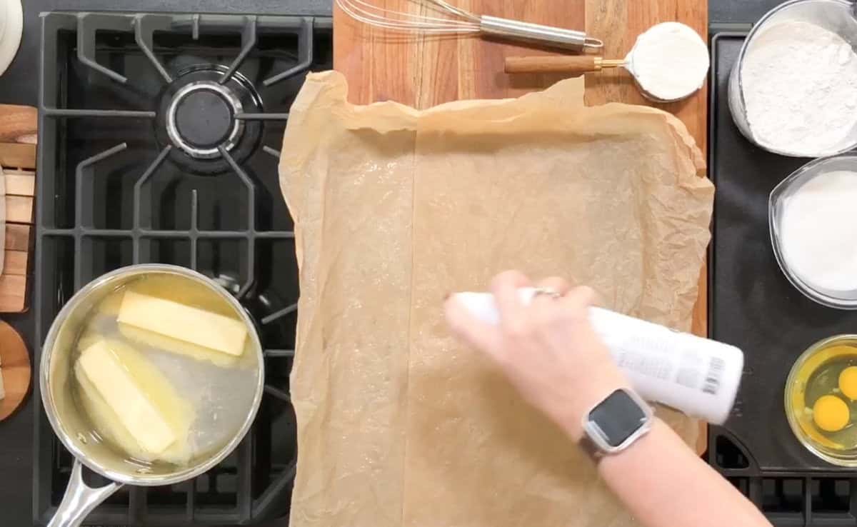 A hand sprays parchment paper on a baking tray with cooking spray. Nearby are a whisk and measuring cup on a wooden board. On the stove, butter is melting in a saucepan. Other ingredients, including eggs and flour, are arranged on the counter.