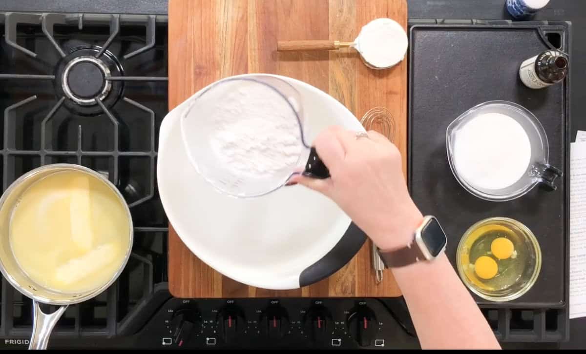 A person is pouring flour into a white bowl on a wooden board. Nearby are ingredients like sugar, eggs in a bowl, a measuring cup, and a whisk. The scene is set on a kitchen stove with a pot of a yellow mixture on the burner.