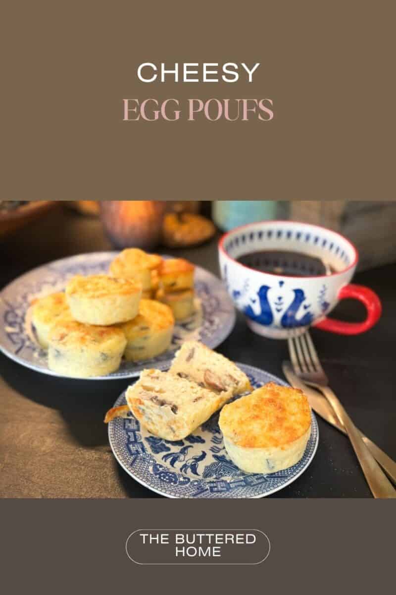 Plates of cheesy egg puffs are displayed, with one puff sliced open to reveal filling. A mug with a blue pattern sits nearby along with a fork. The text "CHEESY EGG POUFS" is at the top, and "THE BUTTERED HOME" is at the bottom.
