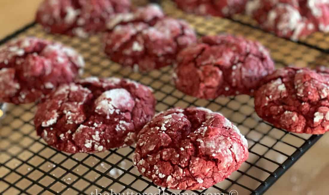 Red velvet crinkle cookies coated with powdered sugar are cooling on a black wire rack. The cookies have a crackled appearance, with a deep red color contrasted by the white sugar. Multiple cookies are visible, evenly spaced on the rack.