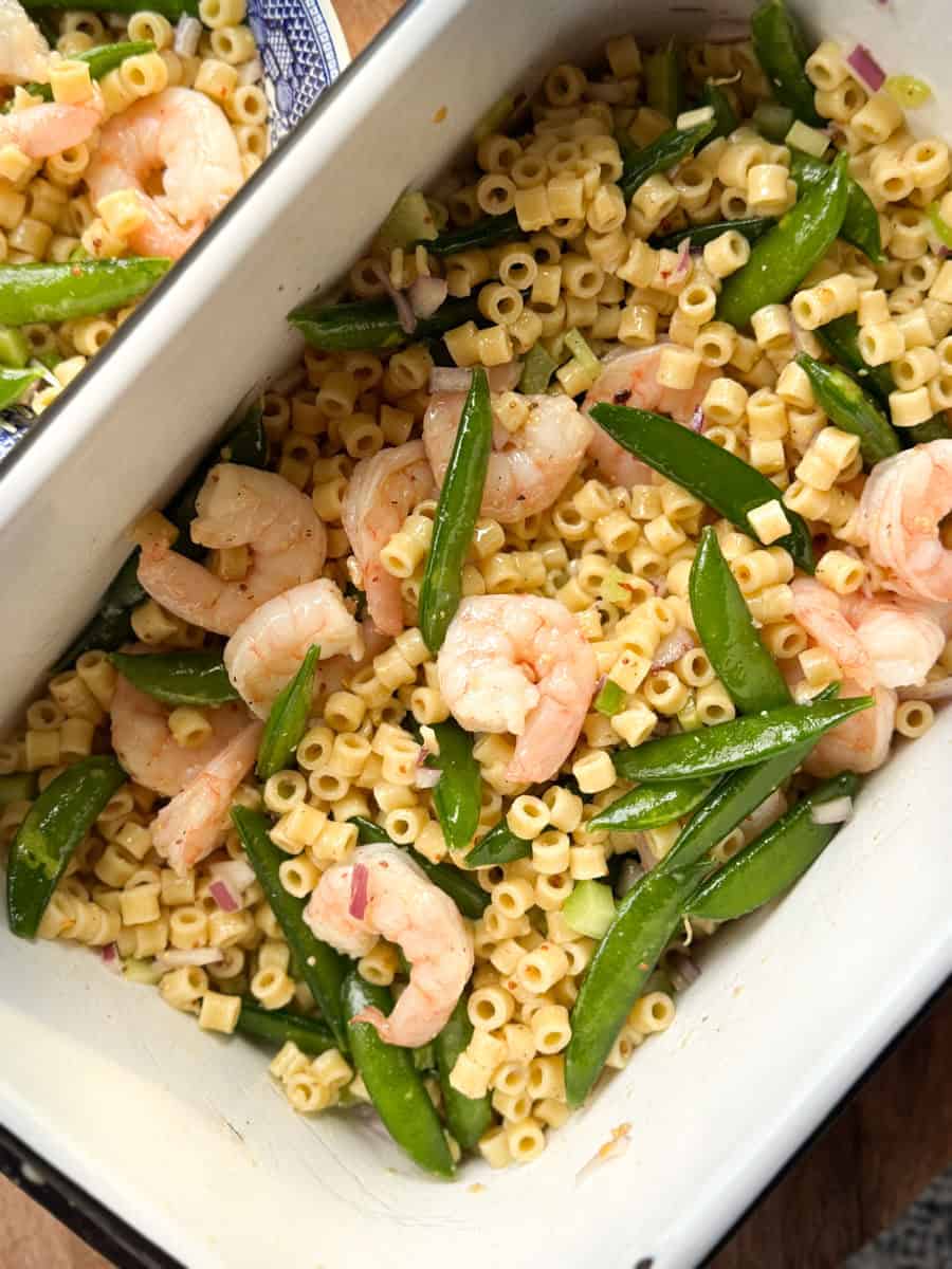A white dish containing pasta salad with shrimp, snap peas, diced red onions, and a creamy dressing. The pasta is mixed with sliced vegetables and shrimp, creating a colorful and appetizing presentation.