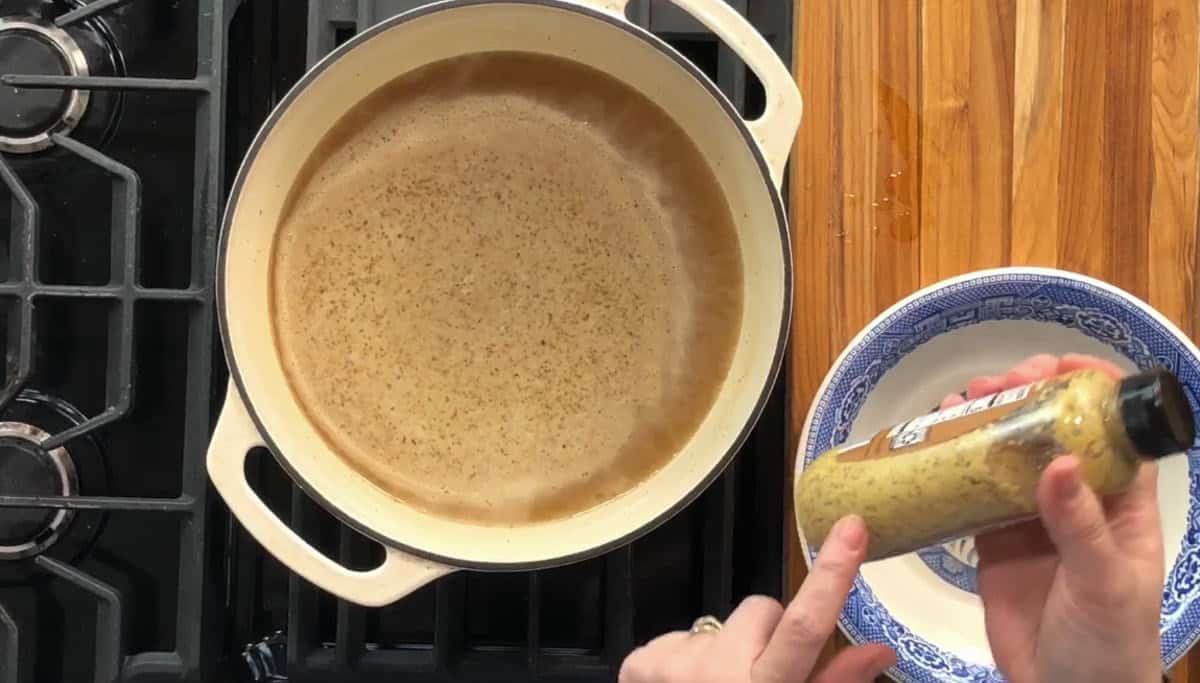 A pot of brown liquid is on a stove next to a blue and white dish containing a yellow substance. A person is holding a bottle of mustard above the bowl, pointing at the label. The stove has black grates and the counter is wooden.