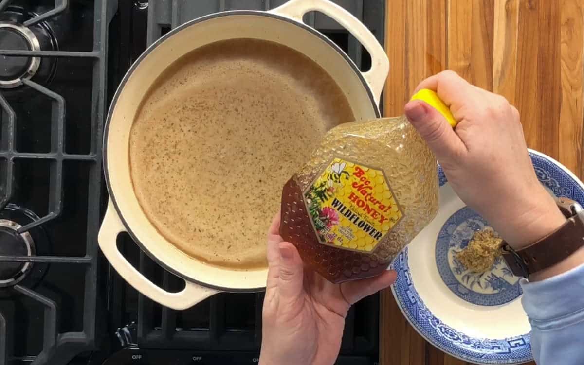 A person is pouring wildflower honey from a plastic bottle into a pot on a stove. The pot contains a brown liquid, possibly a broth or sauce. There is a blue and white bowl with a spoon on a wooden surface beside the stove.