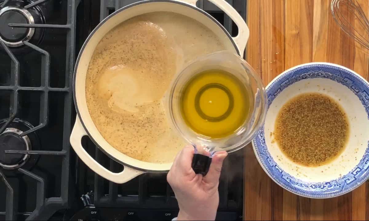 A person is holding a measuring cup with oil above a pot on a stove. The pot contains a frothy, simmering liquid. On a wooden countertop, next to the stove, there's a blue and white bowl with spices. A whisk is nearby.