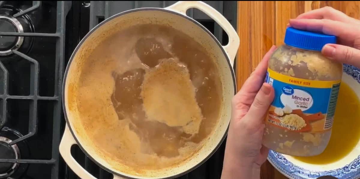 A pot of liquid simmers on a stovetop. To the right, hands hold a jar labeled "Minced Garlic," positioned above a yellow mixing bowl on a wooden surface. The stove has two visible burners.