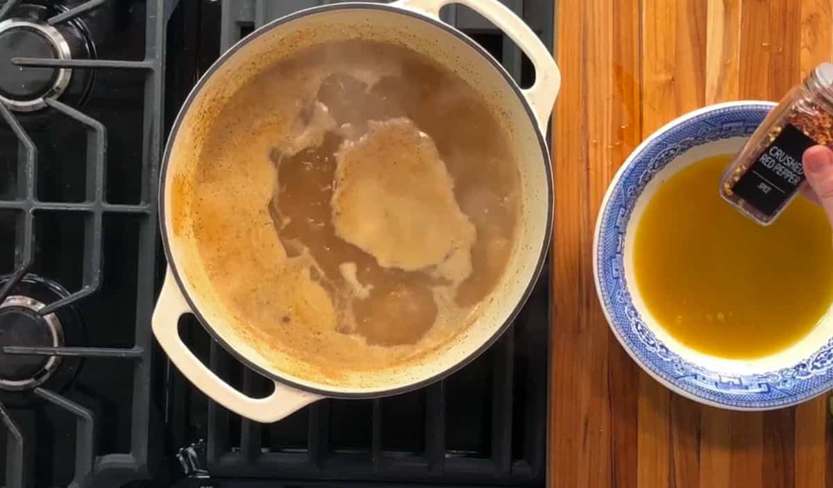 A pot on a black stove with liquid inside, possibly soup or broth, is simmering. To the right, a hand holds a container of crushed red pepper over a blue-and-white bowl containing a yellow liquid on a wooden countertop.