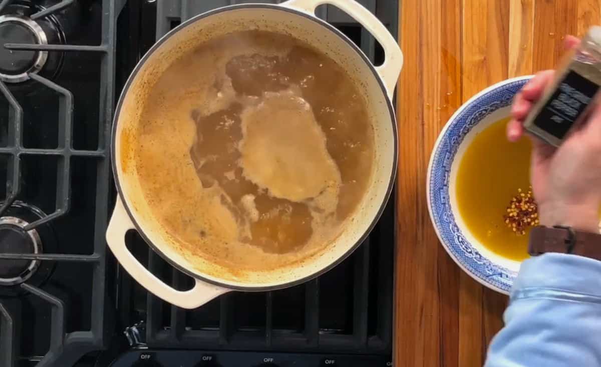 A pot on a stove contains a bubbling liquid, possibly broth or soup. On the right, a person is preparing a mixture with red pepper flakes in a blue and white bowl on a wooden countertop.