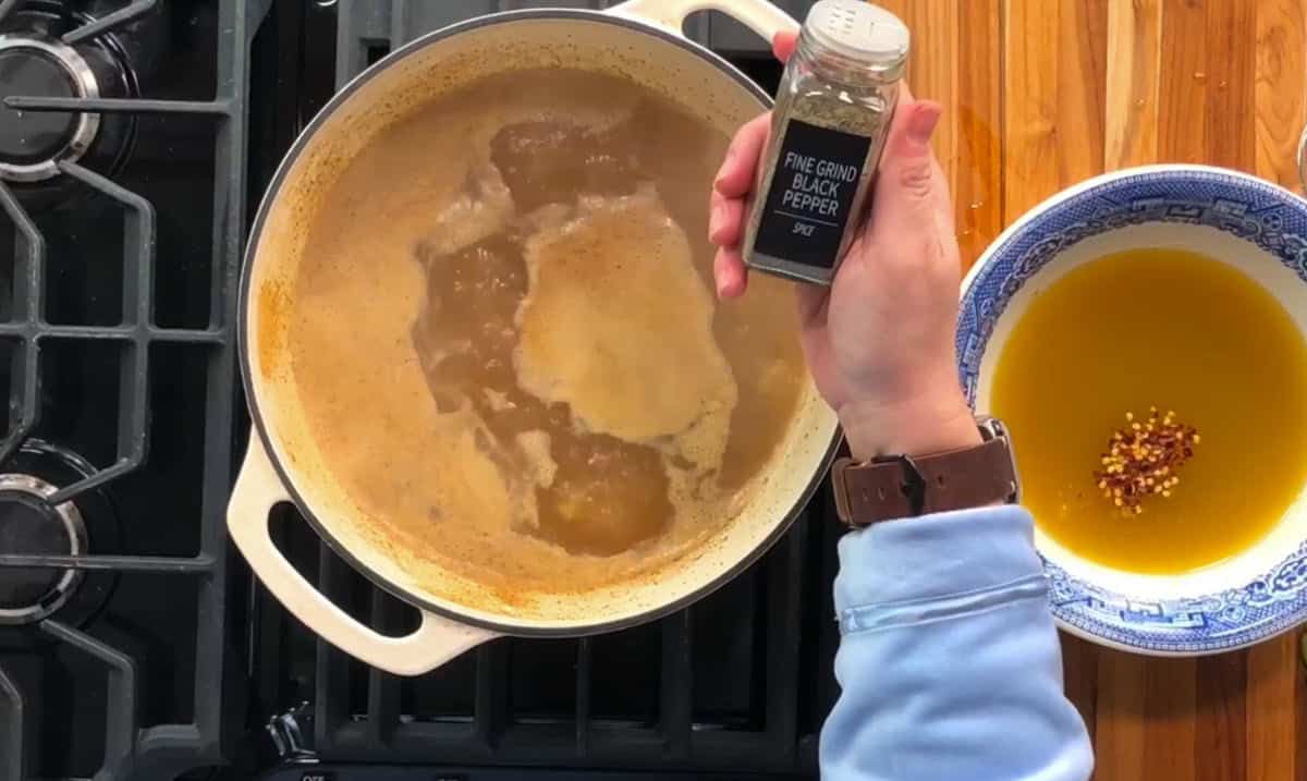 A person holds a jar labeled "Fine Grind Black Pepper" over a pot of simmering liquid on a stove. A bowl containing oil and crushed red pepper flakes sits on a wooden counter nearby.