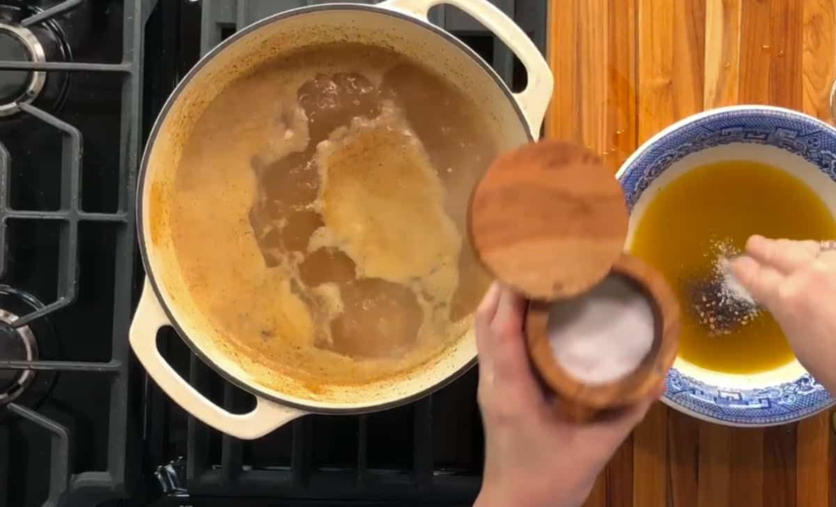 A pot of broth is simmering on a stove. Nearby, a person is adding salt to a bowl filled with a yellow liquid, possibly melted butter. The stove is black, and the counter is wooden.
