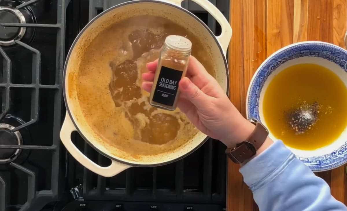 A hand holds a spice jar labeled "Old Bay Seasoning" over a simmering pot on a stovetop. Next to the stove is a wooden countertop with a blue and white bowl containing a yellow liquid and scattered spices.