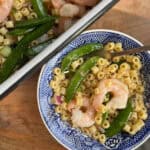 A bowl of pasta salad with shrimp, sugar snap peas, and diced red onion in a blue-patterned dish. A fork rests in the bowl. Next to it is a larger dish with more of the same pasta salad. Both are placed on a wooden surface.