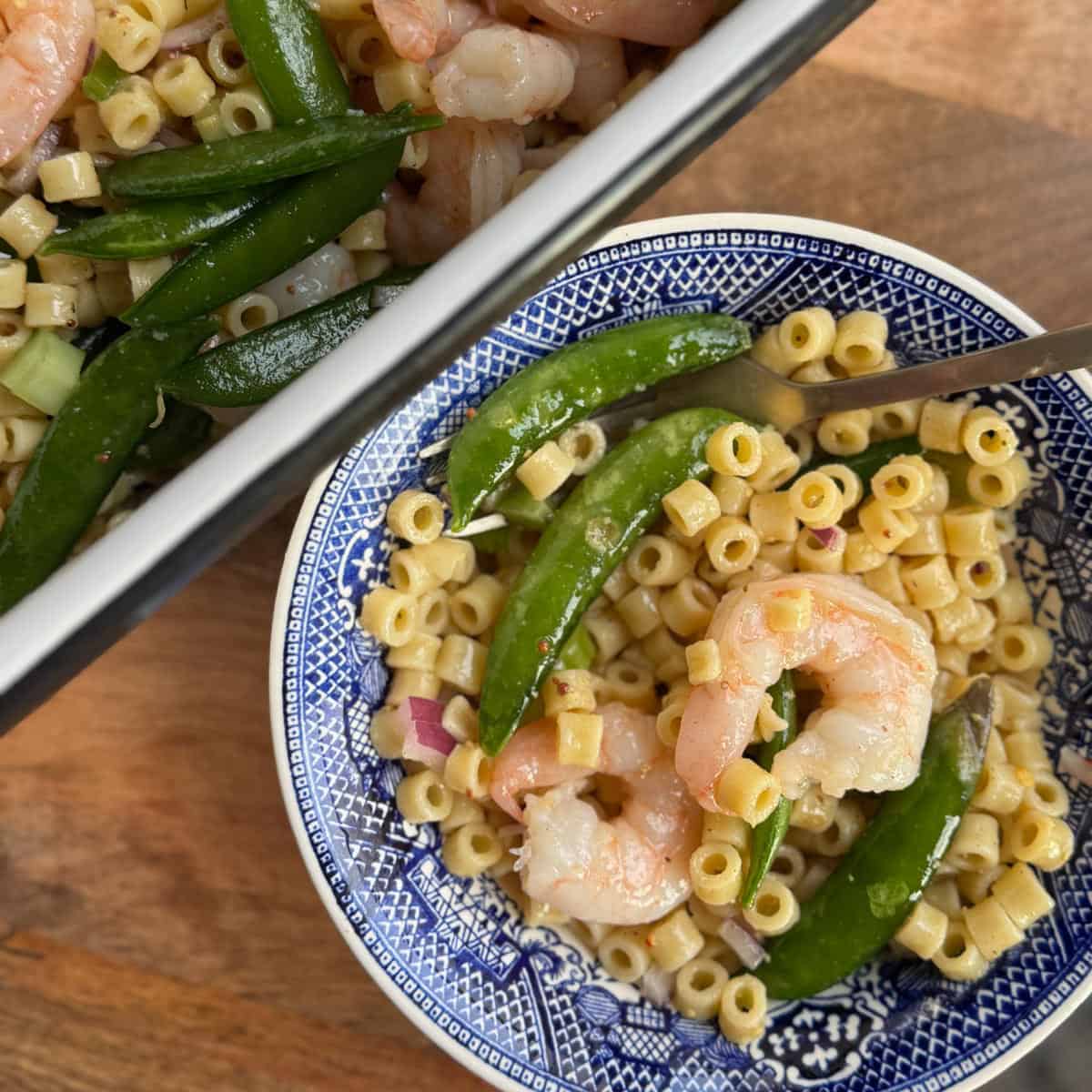 A bowl of pasta salad with shrimp, sugar snap peas, and diced red onion in a blue-patterned dish. A fork rests in the bowl. Next to it is a larger dish with more of the same pasta salad. Both are placed on a wooden surface.