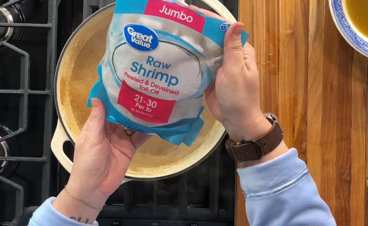 Person holding a bag of jumbo raw shrimp over a pot on a stove. The shrimp are labeled as peeled, deveined, and tail-on, with a quantity of 21-30 per pound. A wooden countertop and part of a blue bowl are visible in the background.