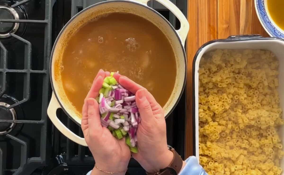 A person holding chopped red onions and green celery over a pot of simmering broth on a stove. To the right, uncooked ditalini pasta is placed in a baking dish on a wooden surface.