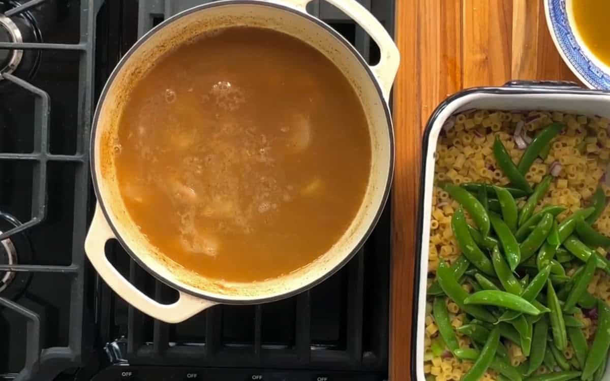 A pot of bubbling brown soup is on a stove. Next to it is a white dish with uncooked pasta and snap peas. A stack of wooden boards is underneath and a glimpse of a blue-rimmed plate is visible on the side.