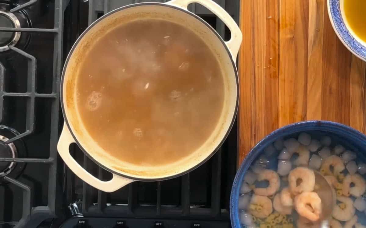 A pot of soup simmers on a stovetop. To the side, a blue bowl contains shrimp submerged in water with ice cubes. The stove is black and the countertop is made of wood.