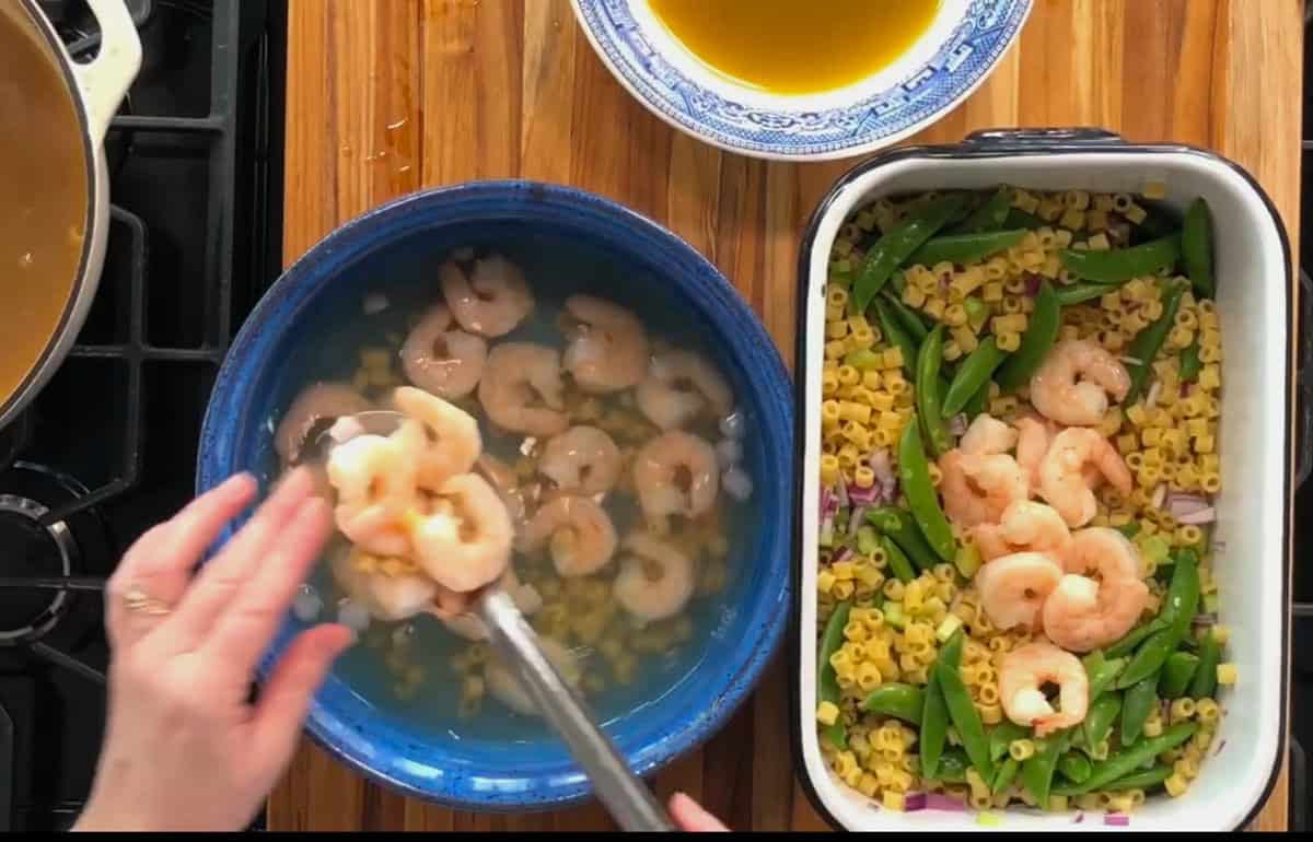 A person is transferring cooked shrimp from a blue bowl with ice water to a baking dish filled with pasta, snap peas, and red onions. A saucepan is on the stove nearby, and a blue-patterned bowl of liquid is on the wooden countertop.