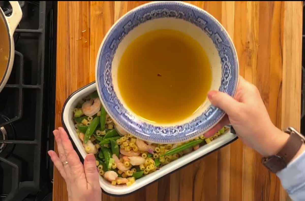 A person pours a yellow liquid from a blue and white bowl over a dish containing pasta, shrimp, and green vegetables on a wooden surface. Part of a stovetop is visible on the left.