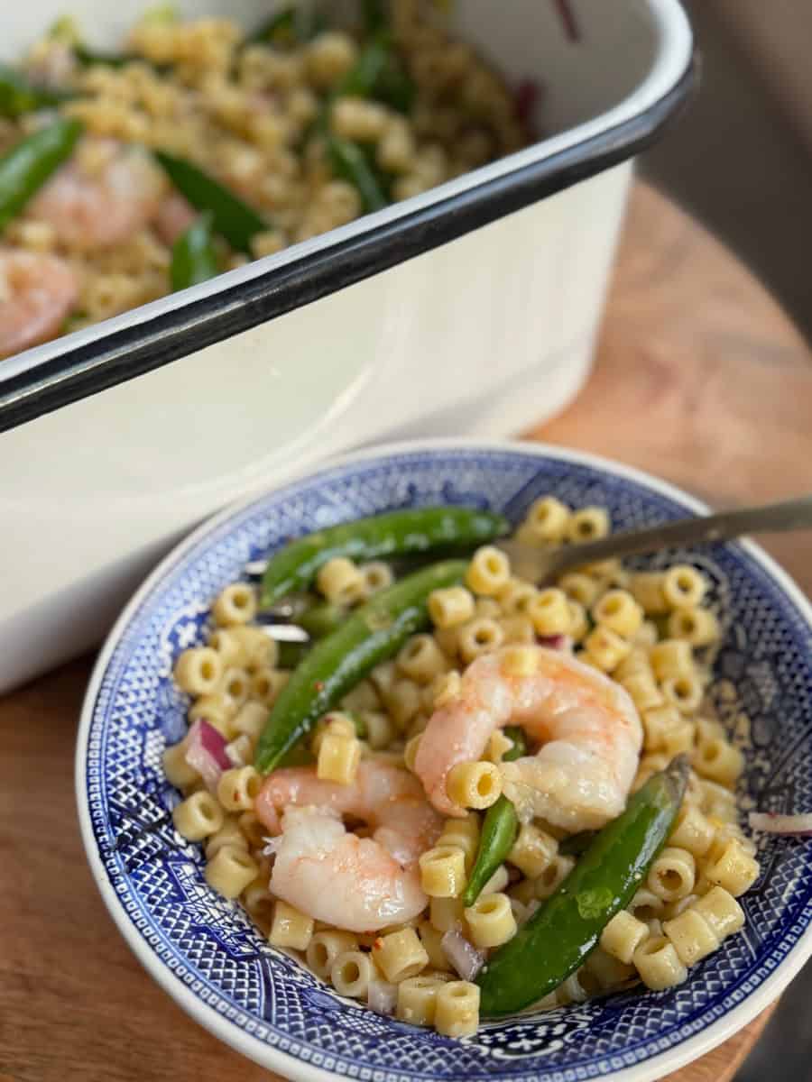 A blue and white bowl filled with pasta rings, shrimp, and snap peas on a wooden surface. A fork is placed in the bowl. In the background, there is a white serving dish with more of the same dish.